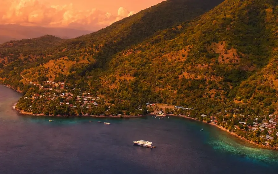 A coastal village on a forested Flores ridge at sunset, with a boat anchored in the bay below