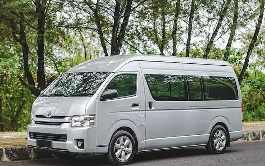 A silver Toyota HiAce van parked on a road beside a stand of trees