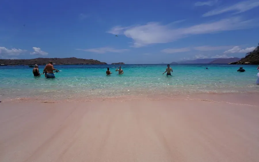 The pink sand of Pink Beach in the foreground with snorkellers standing in clear turquoise shallows