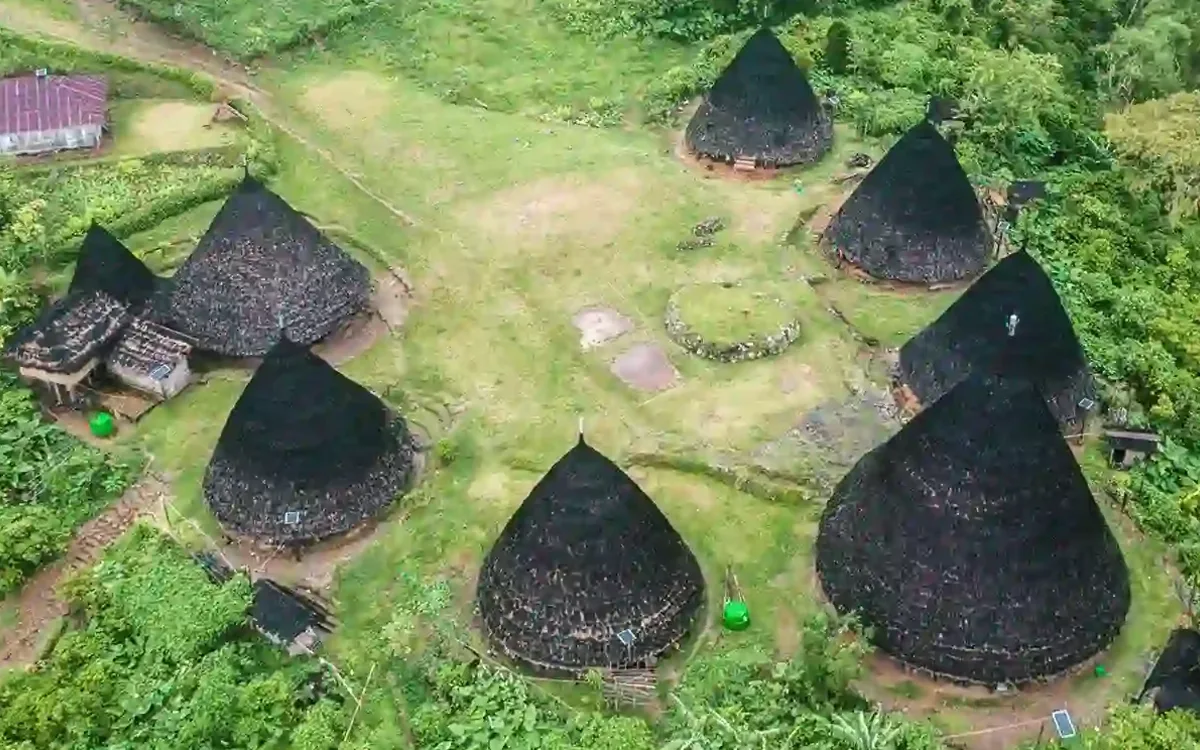 Aerial view of Wae Rebo: seven conical Mbaru Niang houses with dark thatched roofs around a grassed clearing, ringed by forest in the Manggarai highlands