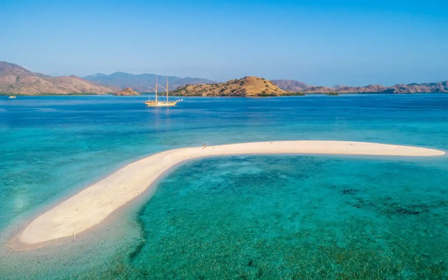 A curved white sandbar at Taka Makassar with a phinisi anchored beyond it