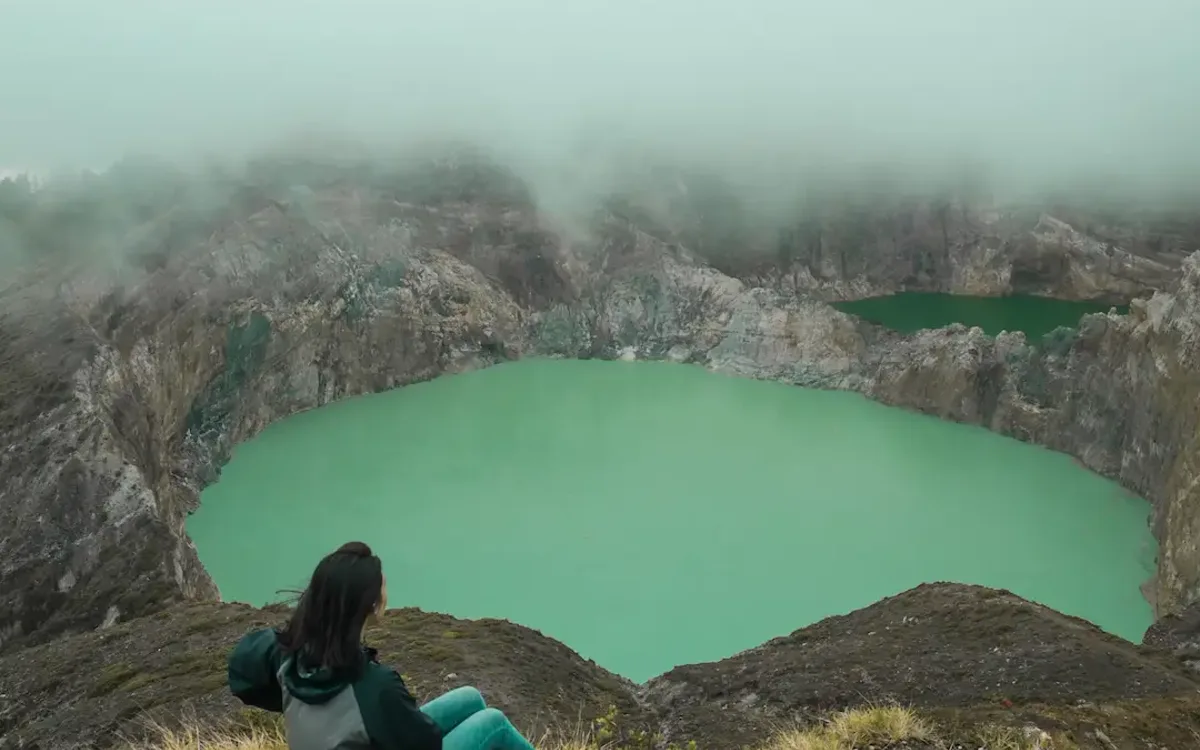 A visitor sitting on the Kelimutu crater rim above a green lake, with mist rolling across the ridge behind
